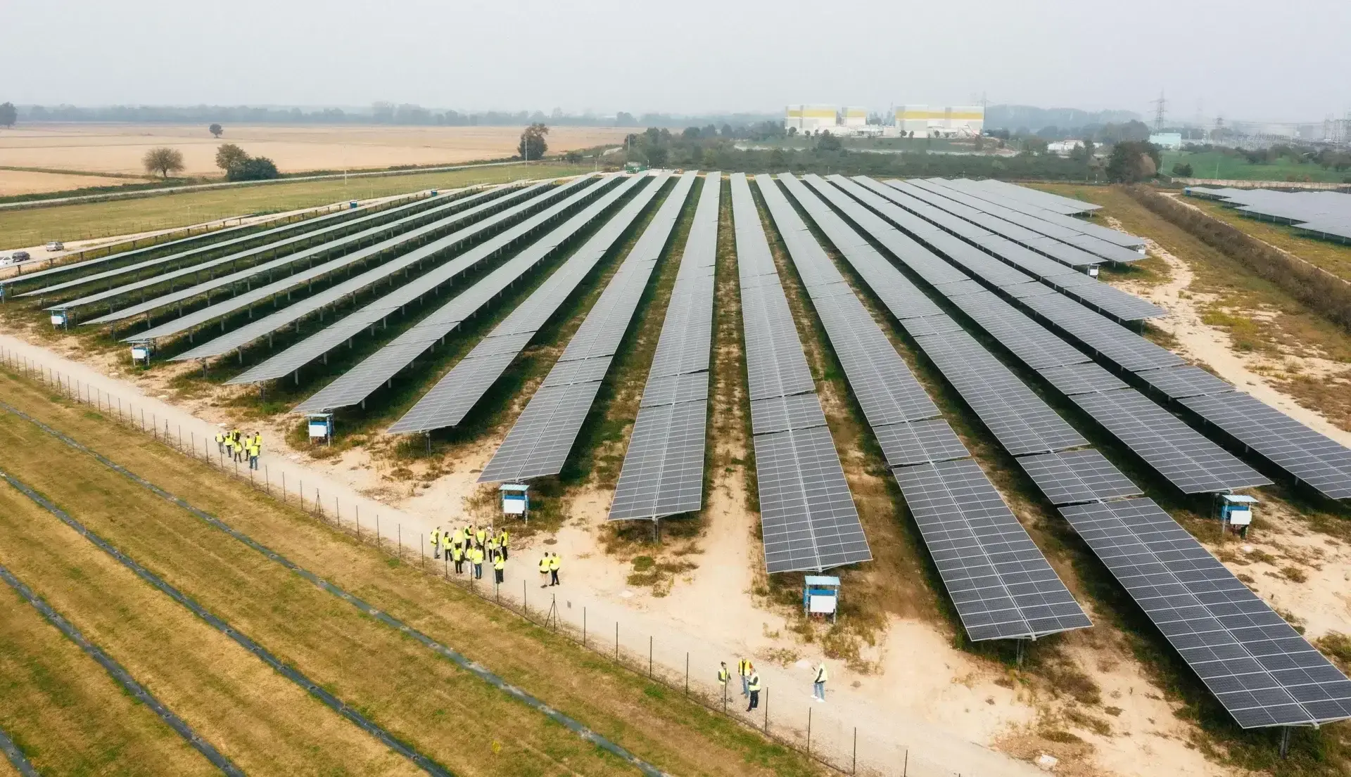 Aerial view of a large solar power installation consisting of multiple rows of photovoltaic panels arranged in a grid pattern across an open field. The solar arrays extend across most of the site, separated by narrow maintenance corridors and patches of grass. A service area with equipment and a few people is visible near the edge of the facility. The image highlights the scale and organization of a utility-scale renewable energy project.