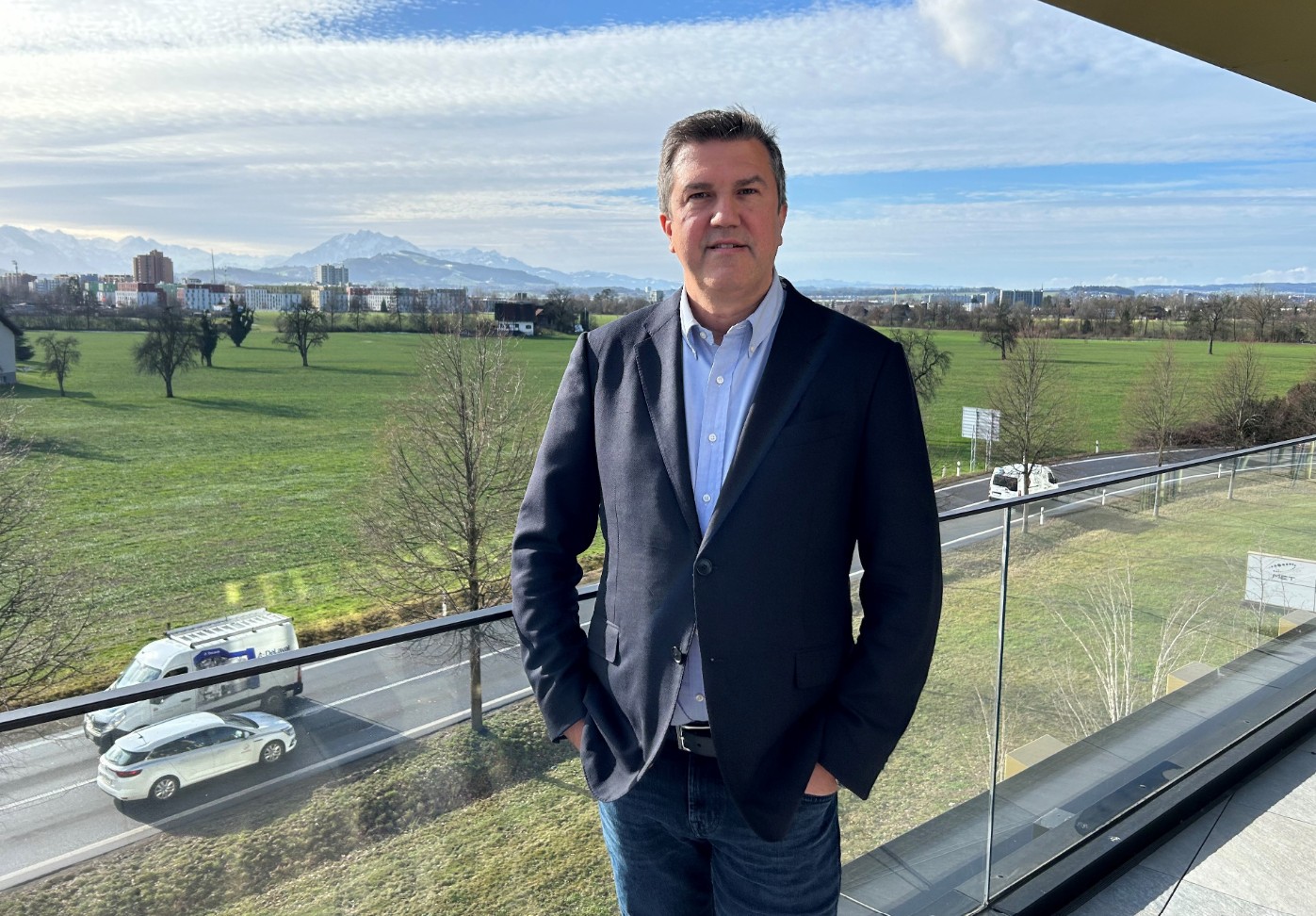 Klaus Reinisch is wearing a dark blazer, light blue button-down shirt, and jeans stands on a balcony overlooking a wide green landscape. In the background, open fields, trees, a roadway with passing vehicles, and distant snow-capped mountains stretch across the horizon under a partly cloudy sky. The glass balcony railing and bright natural daylight create a clear, professional outdoor portrait setting.