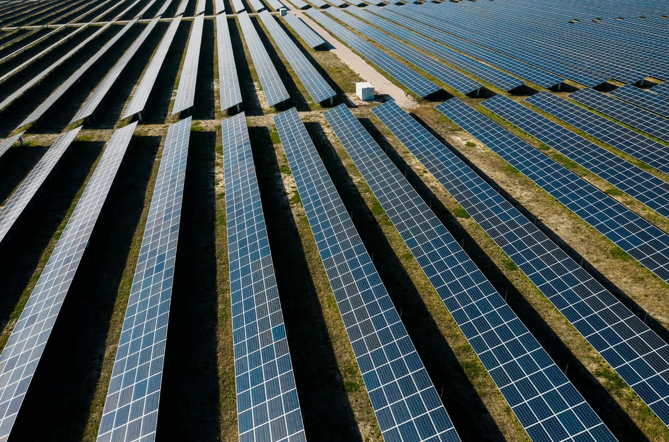 Aerial view of a utility-scale solar power plant with long, parallel rows of photovoltaic panels stretching across a large open site. Dark blue solar modules are arranged in precise geometric patterns, separated by access lanes and strips of grass. A small service structure is visible among the panel arrays. The image highlights large-scale renewable electricity generation infrastructure and the extensive land area used for solar energy production.