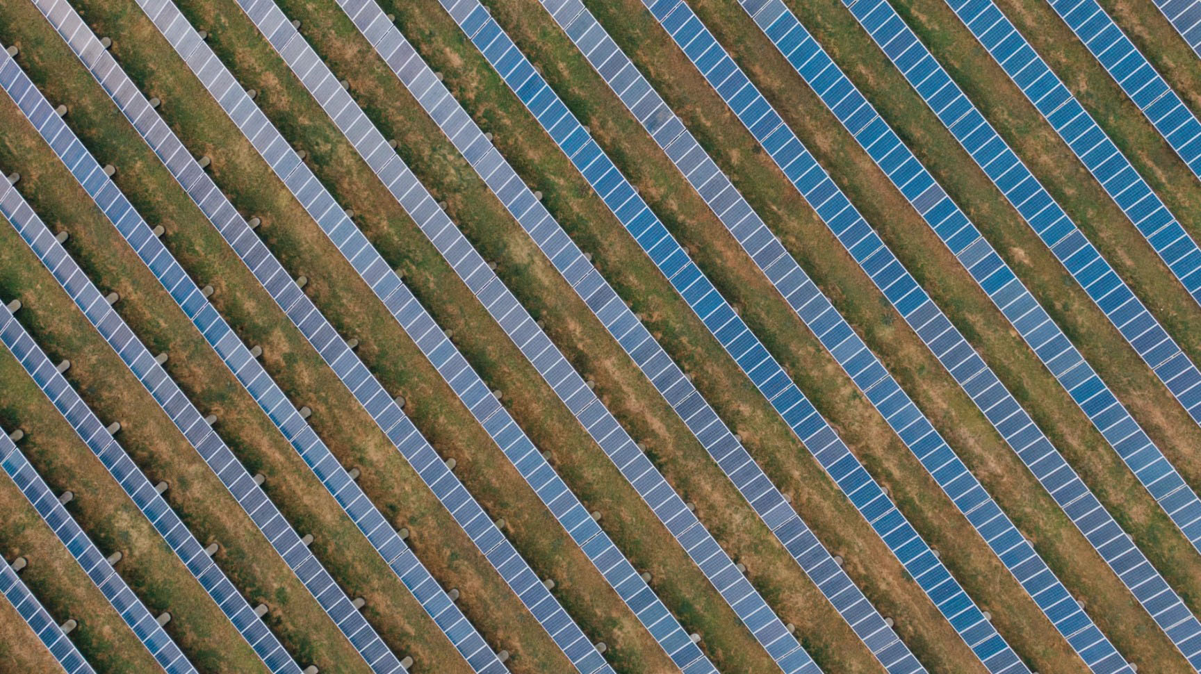 Aerial view of a solar power plant showing long, parallel rows of photovoltaic panels arranged in a precise geometric pattern across open land. The blue solar arrays run diagonally across the image, separated by narrow strips of vegetation and access paths. The overhead perspective emphasizes the scale, symmetry, and orderly layout of the renewable energy facility, illustrating utility-scale solar power generation and sustainable energy infrastructure.